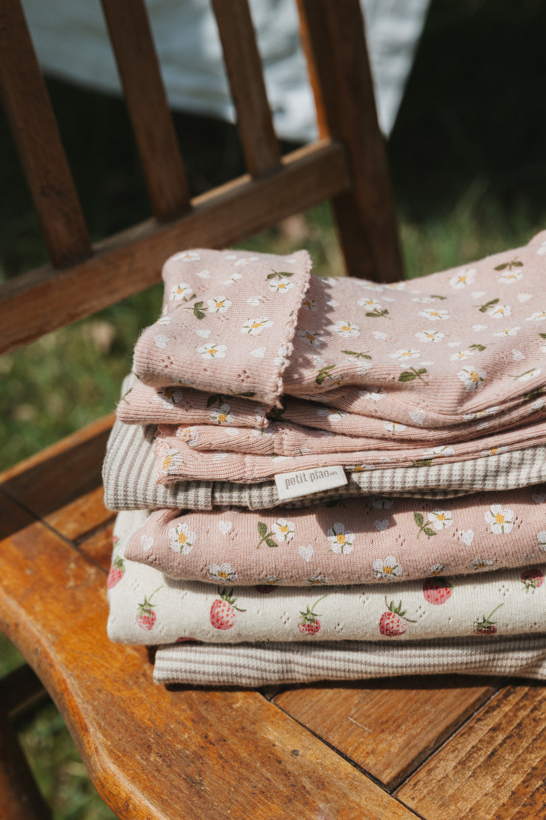 Petit Piao PPDea LS Body Fold NB Fog Strawberry gestapelt mit anderen Hosen und Bodies auf einem Stuhl im Garten 