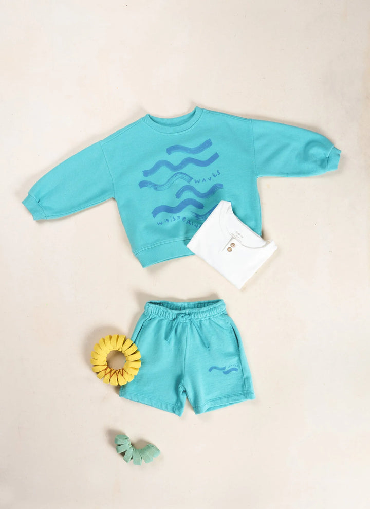 Play Up Fleece Sweater vase auf beigefarbenem Hintergrund liegend kombiniert mit farbgleicher Shorts