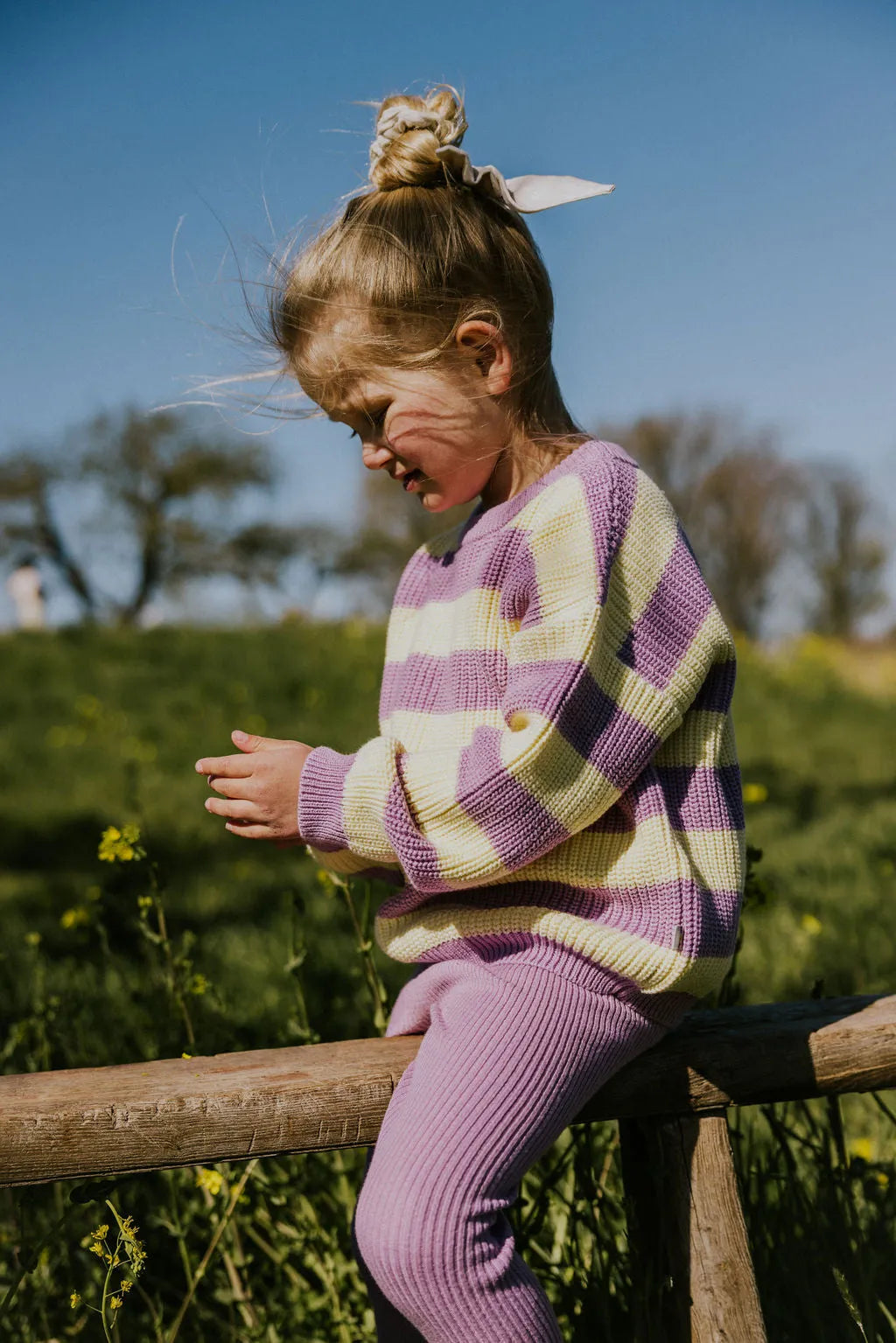 Yuki Stripes Sweater ICE CREAM getragen von einem weiblichen Kleinkind, das auf einem Zaun sitzt, der sich auf einer grünen Wiese befindet, Ansicht von der Seite